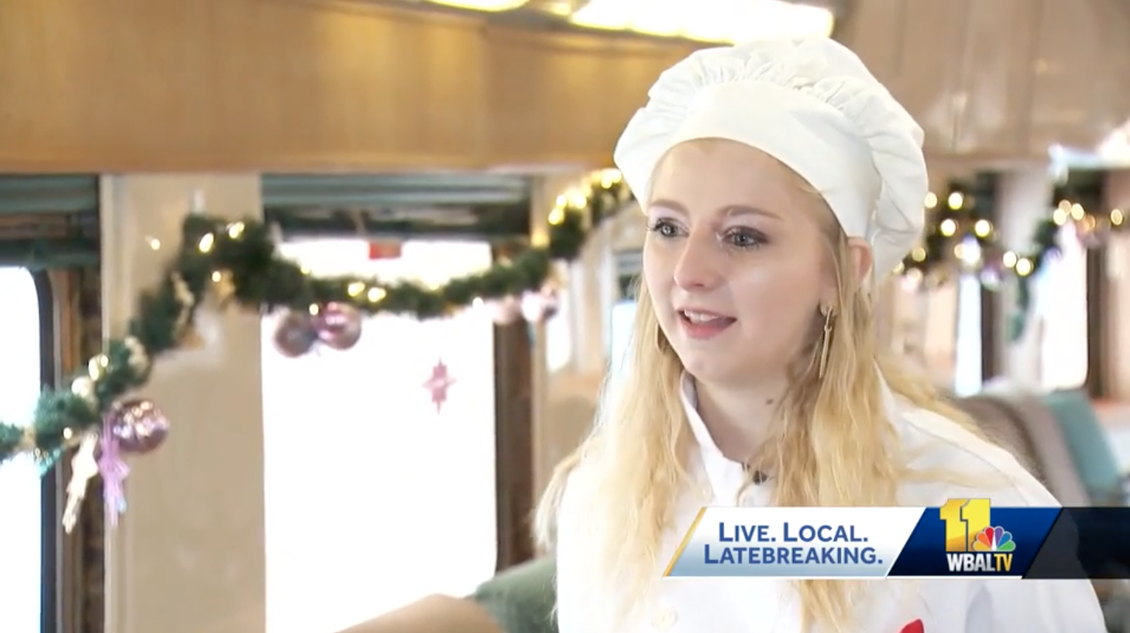 This is an image of a woman standing on a train wearing a chef's jacket and a chef's hat.